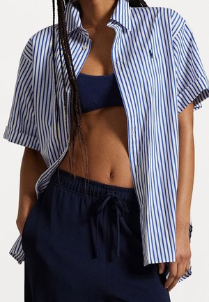 Kvinde iført løs blå og hvid vertikalt stribet kortærmet skjorte over en marineblå crop top og marineblå bukser med snøre.