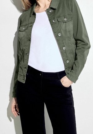 Femme portant une veste boutonnée vert olive par-dessus une chemise blanche et un pantalon noir, une main dans la poche, sur un fond uni.