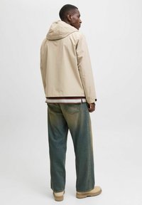 Beige hettejakke i lett materiale med løs passform. Brukt over en stripete skjorte og vide blågrønne jeans. Beige sko fullfører antrekket.