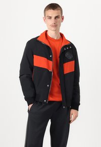 Juodas zip-up švarkas su oranžinėmis juostelėmis, įrengtomis rankogaliais ir apykakle. Viršutinėje kairėje pusėje yra logotipo pleistras. Dėvimas virš oranžinės marškinėlių.