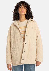 Beige corduroy jacket with diamond quilt pattern, snap buttons, and a collar, layered over a striped sweater, paired with blue jeans.