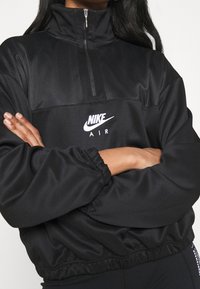 Person som bär en svart Nike Air-pullover med nätaxlar och halvdragkedja, med armarna korsade över bröstet mot en neutral bakgrund.