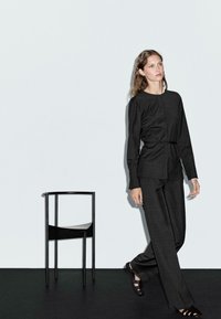 Svart, texturerad outfit som består av en långärmad topp och vida byxor. Inkluderar en figursydd midja med bälte. Svart minimalistisk stol i bakgrunden.