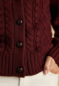 Bordový pletený cardigan s kabelovými vzormi, rebrovaným lemom a veľkými tmavými gombíkmi, s textúrovaným povrchom a priliehavým strihom.