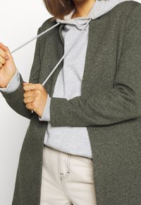 Olijfgroen gebreide cardigan met een relaxte pasvorm, over een lichtgrijze hoodie met een trekkoord. Beige broek met zichtbare stikseldetails.