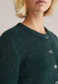 Donkergroene gebreide cardigan, textuurstof, ronde halslijn, twee gouden knopen, zachte uitstraling, gericht op natuurlijke vezels.