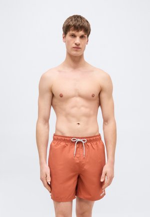 Tânăr cu păr scurt și castaniu, purtând șorturi de baie portocalii cu șnur alb, stând în fața unui fundal simplu de culoare gri deschis.