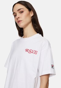 Nuori nainen, jolla on pitkät tummat hiukset, pukeutuneena valkoiseen T-paitaan, jossa on punainen "R.S.V.P." brodeerattu rintaosa ja pieni "F" logo hihassa.