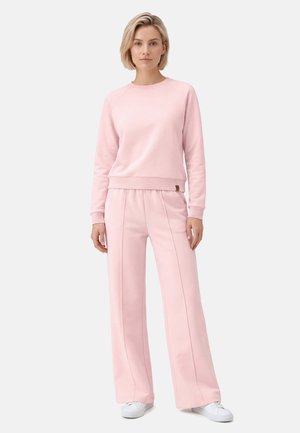 Kobieta stoi ubrana w jasnoróżową bluzę z długim rękawem i szerokie spodnie, białe trampki na jednolitym jasnym tle.