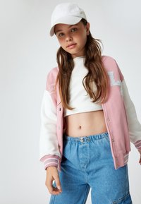Roze en witte varsityjack met geribbelde manchetten, een cropped witte top en blauwe denim broek. Bevat geborduurde details en drukknoopsluitingen.