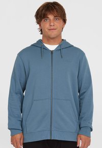 Blue zip-up hoodie made of soft fabric, featuring a hood, two front pockets, and a small logo on the left chest. Smooth texture.