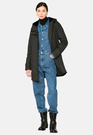 Zwarte parka-jas met een gevoerde capuchon van bont, gedragen over een blauwe denim jumpsuit. Model draagt zwarte enkelboots. Effen witte achtergrond.