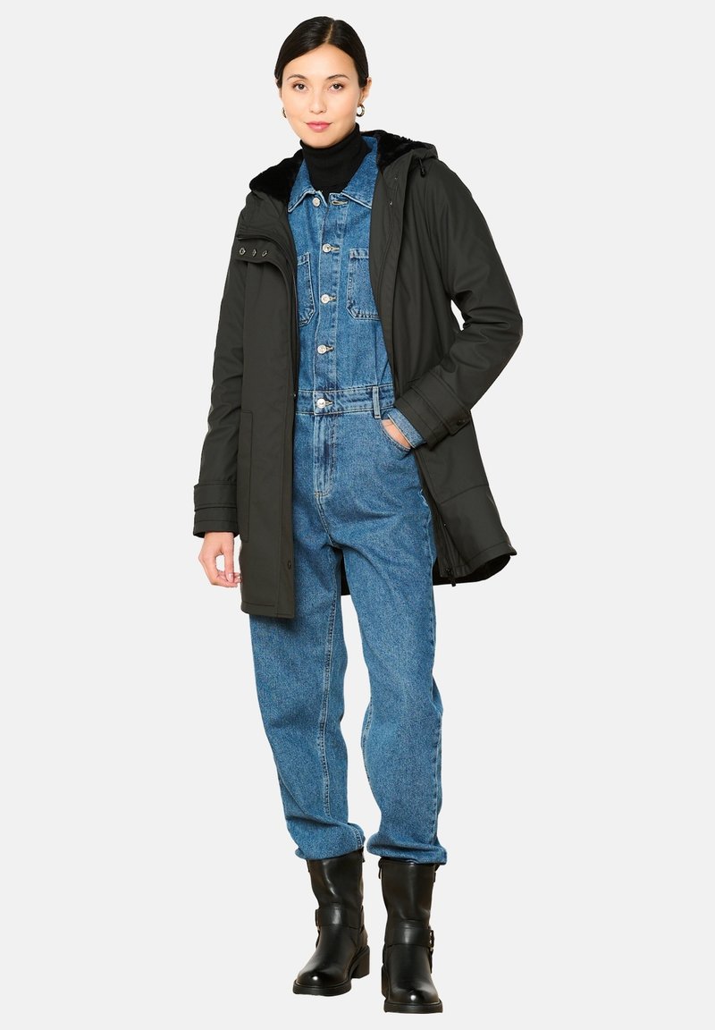 Juodas parka tipo švarkas su kailiniu kapucėje, uždėtas ant mėlynos džinsinės šokinėjimo aprangos. Modelis dėvi juodas kulkšnies ilgio batus. Paprasta balta fono.