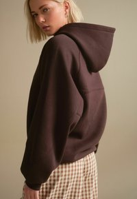 Sweatshirt à capuche marron avec une coupe décontractée, des épaules tombantes et des poignets côtelés, associé à un pantalon à carreaux beige. Tissu lisse et uni.