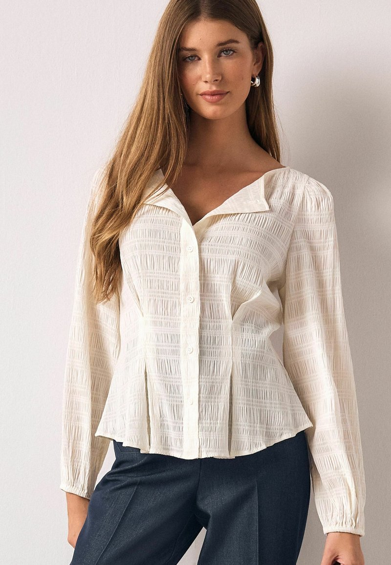 Hvid skjortebluse med knapper, tekstureret mønster, krave med fold og lange ærmer. Kombineret med mørke bukser.