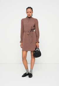 Femme debout, portant une robe longue à carreaux bruns, des chaussettes grises, des mocassins noirs, et tenant un petit sac à main noir.