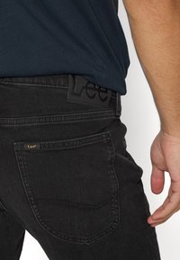 Mustat farkut, joissa on takatasku ja mustasta nahasta valmistettu logo "Lee". Kangas on sileäpintaista ja siinä on selkeä ommel.