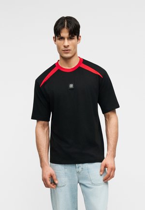 Jeune homme se tenant face à l'avant, portant un T-shirt noir avec des accents rouges aux épaules et un jean bleu clair, devant un fond gris uni.