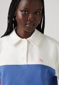 Balta un zila polo krekls ar apkakles izgriezumu, trīs pogām un rozā izsistiem logos uz krūtīm. Strukturēts audums.