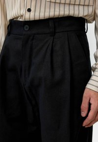Zwarte geplooide broeken van een gestructureerde stof, met een tailleband en twee zijzakken. Gecombineerd met een gestreept beige shirt.