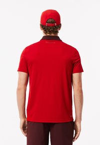 Röd polo skjorta med korta ärmar och en mörk krage. Har en Lacoste etikett på baksidan. Röd keps med justerbart band.