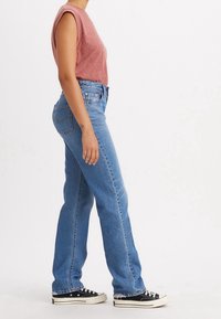Top sin mangas de color rojo claro combinado con jeans rectos azules, acentuados con dobladillos deshilachados. Zapatillas negras completan el conjunto.
