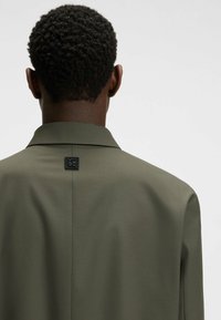 Olivgrön blazer med en jämn, matt textur. Har en klassisk krage, ryggslits och en liten logodetalj nära halslinjen.