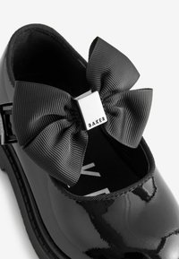 Schwarzer Lackleder-Mary-Jane-Schuh mit einer breiten, strukturierten schwarzen Schleife und einem silbernen quadratischen Akzent mit der Aufschrift "BAKER". Die Innensohle zeigt das Logo.