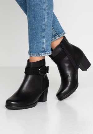 Sorte ankelstøvler i læder med chunky hæl, elastisk rem med cirkulært metalspænde og teksturerede detaljer på hælen.