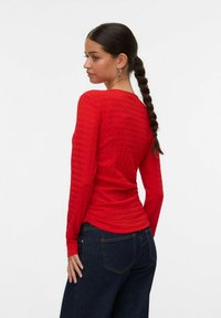 Kvinne med langt flettehår iført en strukturert rød langermet topp og mørkeblå jeans, vendt mot siden mot en ensfarget hvit bakgrunn.