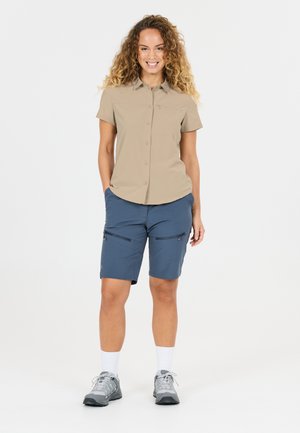 Frau steht lächelnd da, trägt ein beige Kurzarm-Hemd mit Knöpfen, blaue knielange Shorts mit Reißverschlusstaschen, weiße Socken und graue Turnschuhe.