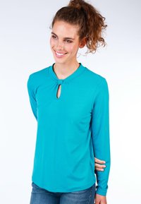 Langärmliges türkisfarbenes Oberteil mit einem Ausschnitt im Schlüssel-Schloss-Stil und weicher Textur, kombiniert mit blauen Jeans.