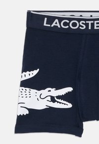 Tengerész kék pamut boxer alsónadrág, nagyméretű fehér krokodil grafikával és kontrasztos fekete derékpánttal, amelyen a "LACOSTE" felirat található.