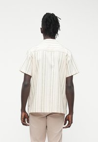 Vyras su juodais dredais, dėvintis trumpomis rankovėmis kremine marškinėliai su plonomis vertikaliomis juostelėmis ir smėlio spalvos kelnes, parodytas iš nugaros ant balto fono.