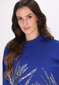 Kék kötött pulóver, magas nyakkivágással és az elején levélre emlékeztető dekoratív ezüst flitteres mintával. Puha tapintású.