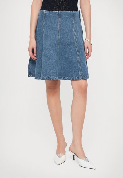 Jupe en denim en bleu moyen avec des coutures visibles, un design évasé et une coupe mi-longue. Associée à des mules blanches à bouts pointus.