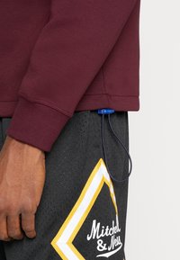 Persona vestida con un sudadera color burdeos y pantalones cortos negros de Mitchell & Ness, con un toggle de plástico azul adjunto al dobladillo de la sudadera.