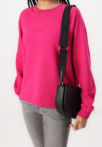 Fuksjowa bluza z ribbowym dekoltem i długim rękawem, w połączeniu z czarną torbą crossbody z teksturowanym paskiem i zakrzywionym kształtem.