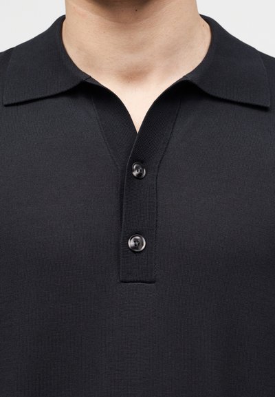 Κοντινό πλάνο ενός ατόμου που φοράει μαύρο πλεκτό πόλο με δύο μαύρα κουμπιά και ανοιχτό γιακά.