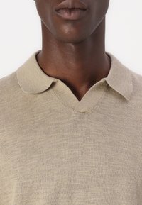 Polo de punto en color beige con cuello en V y corto, en punta. La textura parece suave y ligera, con un patrón sutil y uniforme.