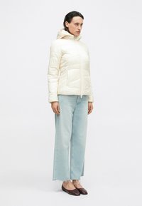 Giubbotto imbottito di un delicato crema con cappuccio, caratterizzato da una texture trapuntata, zip frontale, abbinato a jeans a gamba ampia di un lieve blu e scarpe slip-on scure.