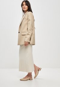 Beige räätälöity bleiseri, jossa on yksi nappi, yhdistettynä kermaiseen midi-hameeseen. Lyhyet beige slingback-kengät, joissa on matala metallikoristeinen korko.