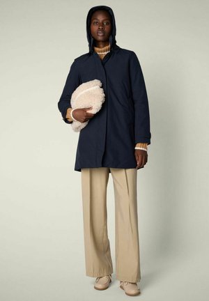 Donna con cappotto blu navy con cappuccio, pantaloni beige a gamba larga e scarpe marroni, che tiene in mano una pochette pelosa color crema su uno sfondo semplice.