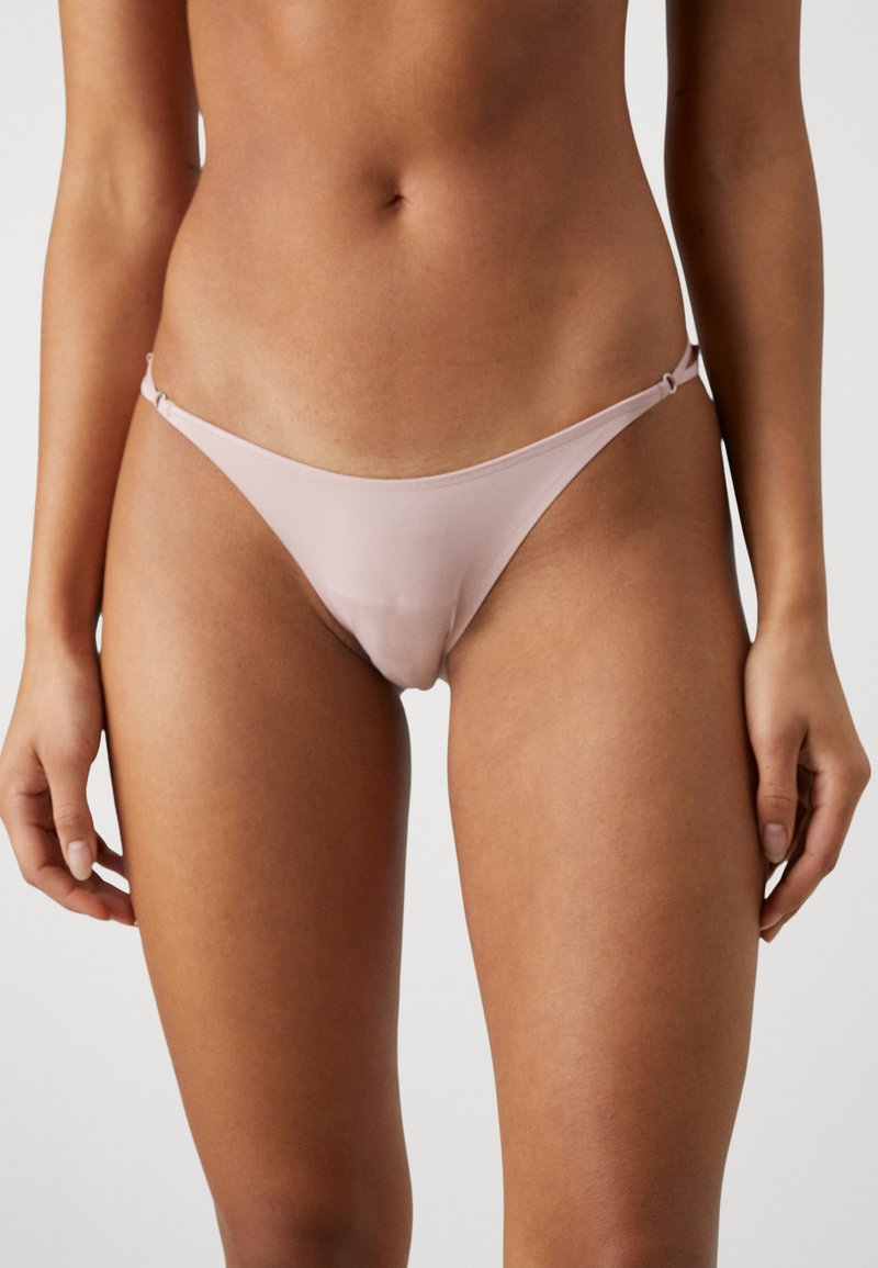 Thong roz deschis cu bretele ajustabile, având o textură netedă și un design minimalist, conceput pentru confort și potrivire.