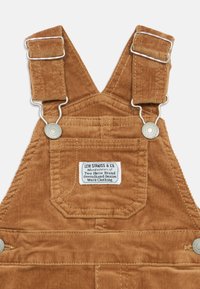 Salopete de algodão castanho com fechos de metal, apresentando uma etiqueta de marca e uma textura distinta de nervuras verticais. Design durável e funcional.