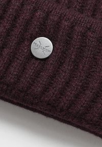 Tamno ljubičasta pletiva s malom okruglom srebrnom metalnom oznakom ugraviranom s "Pearlwood" i grafikom igle.