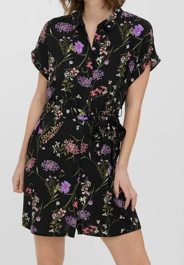 Femme portant une robe noire ceinturée à manches courtes avec des motifs floraux multicolores et un col à boutons.