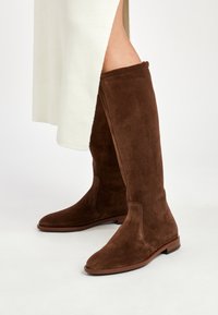Botas de ante marrón hasta la rodilla con una textura suave, puntera redondeada y tacón plano. La caña de la bota presenta un diseño limpio y sin costuras.