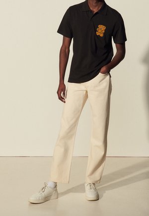 Mężczyzna w czarnym polo z naszywką misia, kremowych spodniach, białych skarpetkach i białych trampkach, stojący z jedną ręką w kieszeni.