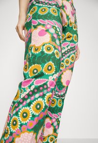 Primo piano di una persona che indossa pantaloni a fantasia floreale colorata con fiori verdi, rosa, gialli e bianchi, in piedi su uno sfondo semplice.